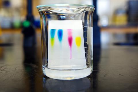 An image showing a beaker containing a thin layer chromatography plate with colourful spots on it