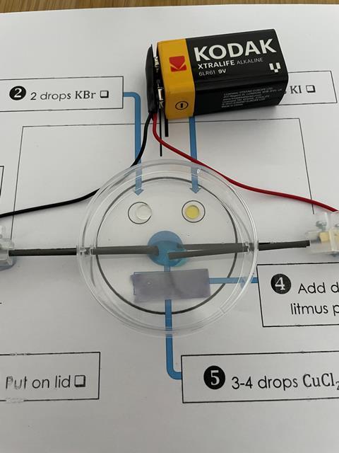 Showing the set up of this experiment. A Petri dish is placed on top of a print out of the integrated instructions which acts as a guide to place the litmus paper, electrodes and drops of solution. The lid is on the petri dish a 9 V battery is attached