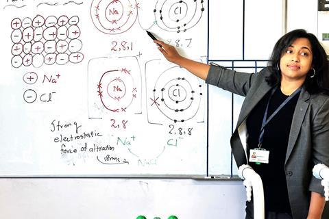 Suriya teaching a class at the whiteboard. The lesson is on ionic bonding and she is showing using electron shell diagrams how sodium donates an electron to chlorine to make sodium chloride.