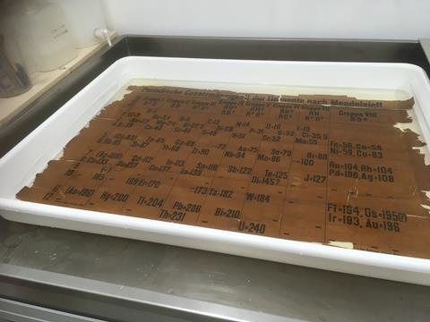 A metal workbench holds a white shallow tray, filled with a liquid and an old document, the oldest classroom example of the periodic table