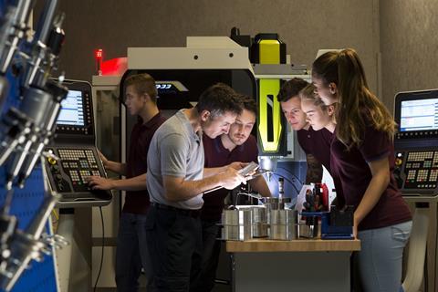 Mature male with a small group of male and females, he is using a bore micrometer and a vernier to measure. Standing in a confined dim lit area surrounded by machinery.