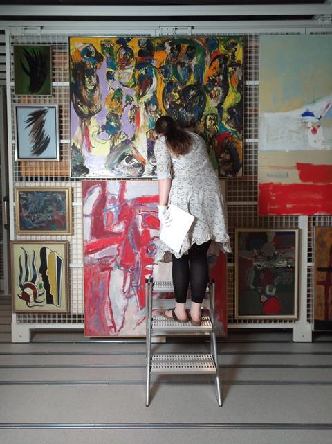 An image showing Ida Antonia Tank Bronken, from behind, on a stepladder examining one painting among many on the wall