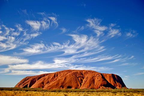 Uluru in Australia - a large orange-coloured rocky hill in a dry landscape with a cloudy blue sky