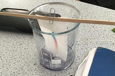 A beaker with liquid soaking up a piece of paper spreading out some ink.
