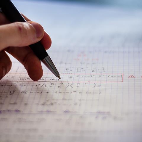 Hand holding a pen over a page of equations, out of focus
