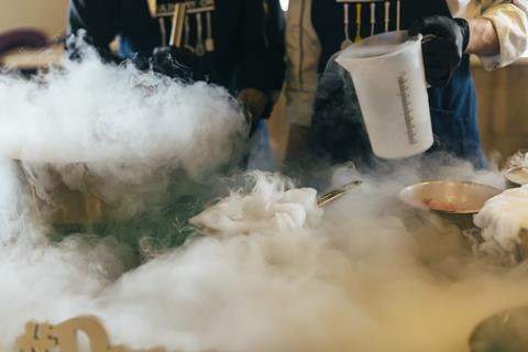 A photo of two chefs with pans and smoke while making ice cream with liquid nitrogen