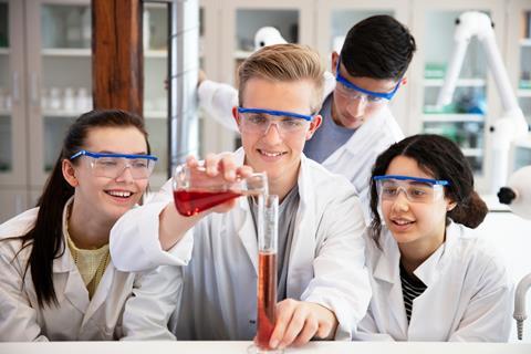 Four high school students in a practical chemistry lesson wearing goggles and lab coats