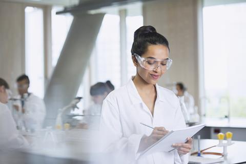 An image showing a female student writing notes in a notebook she's holding; she is wearing a lab coat and lab spectacles, and the background is a school science laboratory