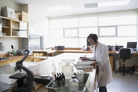 A picture showing a female technician wearing a white lab coat in a school laboratory with a textbook, microscope and fume cupboard visible