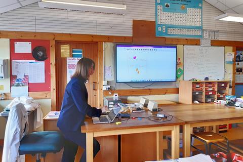 A teacher at the front of a science classroom draws on a laptop screen connected to a large display screen
