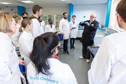 A class of chemistry teachers in white lab coats watch a demonstration by a teacher trainer in a school lab