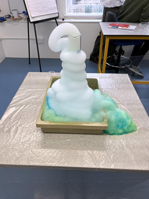 An elephant toothpaste experiment showing a green and white foam coming out of a large glass flask in a classroom setting.