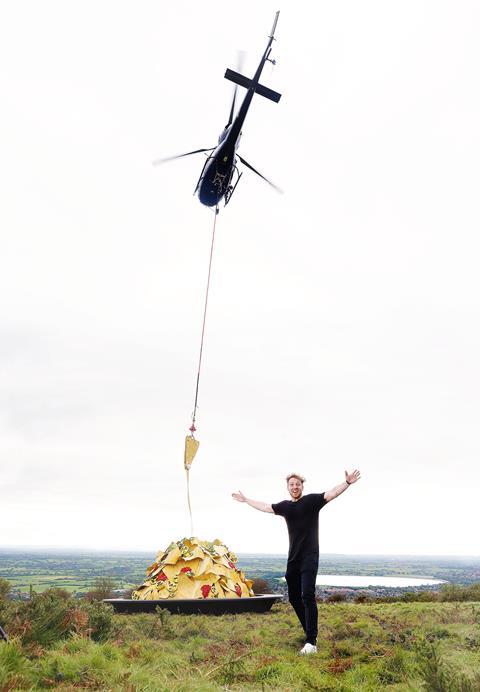 A happy man standing in front of a flying helicopter which is pulling a long string of melted cheese from a mound of giant nachos