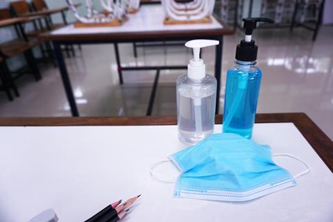 An image showing a medical mask, bottles of hand sanitzer and pencils on a desk in a school classroom.