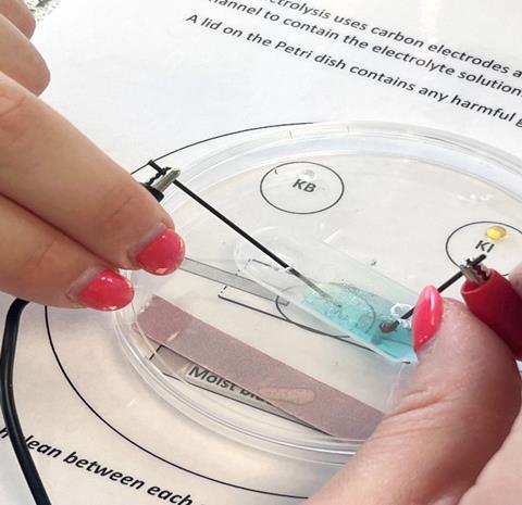 Close up of hands performing a microscale electrolysis experiment in a pertri dish with a blue solution and two anodes joined up to crocodile clips
