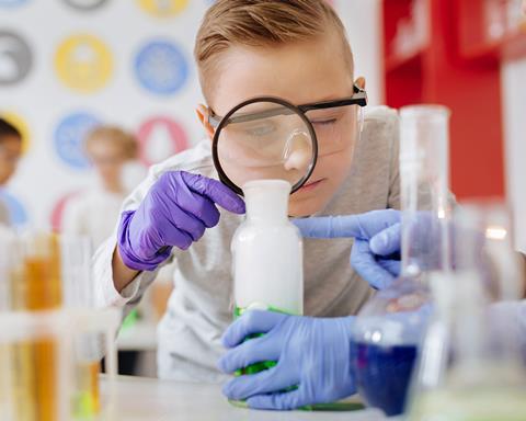 A picture showing a student with a visual impairment wearing safety glasses and using a magnifier; someone is holding the beaker he is looking at