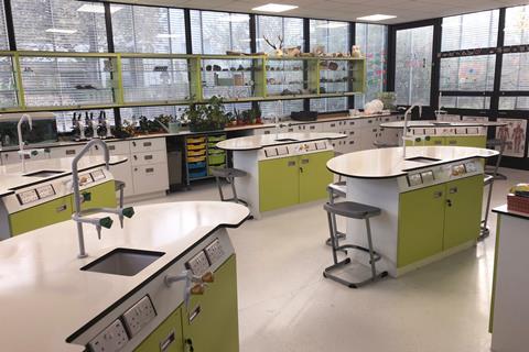 The inside of a school science lab with bean-shaped student desks that have a sink and taps, gas taps, a cupboard and sockets. Along one side of the room is a large glass window with display cupboards in front holding lab glassware and scientific objects.