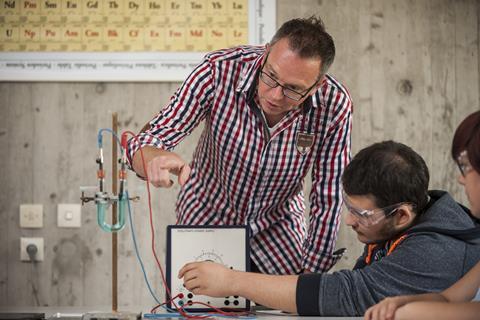 Periodic table on the background wall, teacher wearing glasses demonstrating pointing to electrochemistry with two seated boys wearing safety glasses