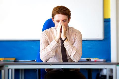 An image showing a stressed teacher, sat at a desk, with hands in front of face. Blue walls, whiteboard in background