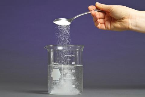 A hand pouring a spoon of sugar into a beaker of water