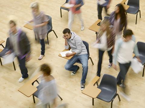 An image showing college students moving around man at desk in a classroom