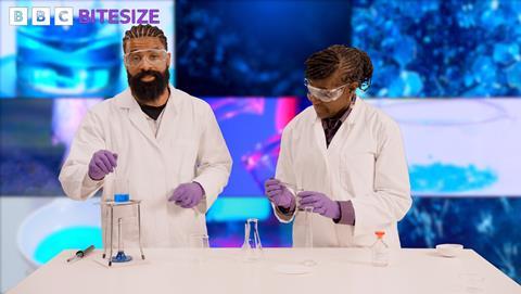Two people in lab coats, safety goggles and latex gloves using a bunsen burner and other lab equipment