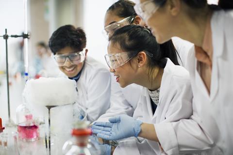 Four students wearing lab coats, safety goggles and gloves work at a lab bench