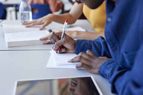 A photo of a student taking notes using a pen and notepad, with another student in the background reading a textbook