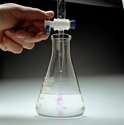A close up photo of titration equipment with a liquid dripping into a conical flask turning purple