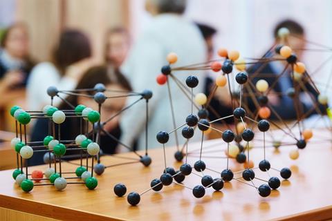 Models of different types of chemical bonding including diamond and salt on a table in a classroom. Behind a teacher speaks to the high school students.