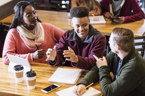 Three students sitting at a table with a tablet, pens, paper, having a discussion