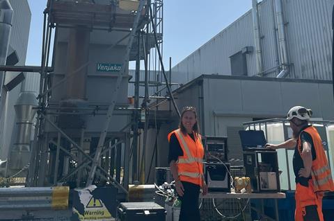 Emma is at a plant wearing a fluorescent orange piece of outer clothing.