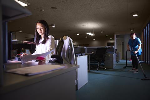 Woman working late at her desk, cleaner vacuuming in background