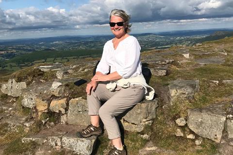 Jill is sitting on some rocks, wearing sunglasses and smiling. There are hills in the background.