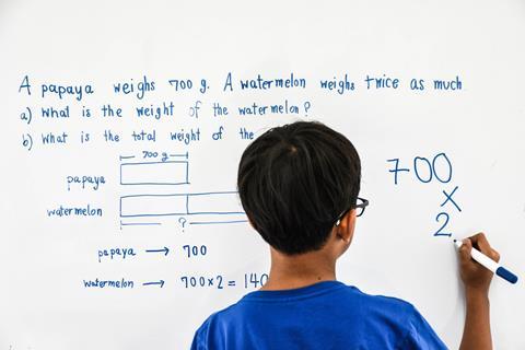 An image showing a child (wearing a blue T-shirt) solving a maths problem using the bar model on a whiteboard