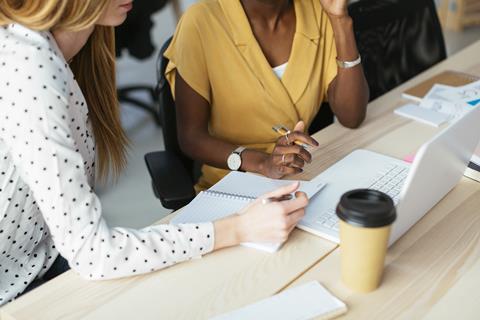 Two people sit together at a long desk looking at a laptop, with an open notebook and pens to take notes