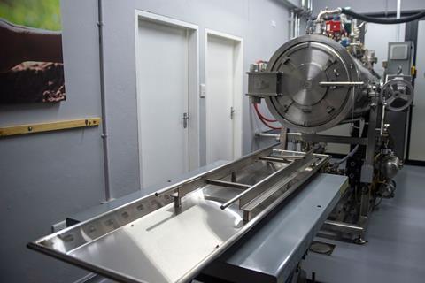 A shiny metal tube large enough to take a human body with a round metal door at one end and machinery on the top including a pipe for liquids. In front is a table with a long metallic tray on.