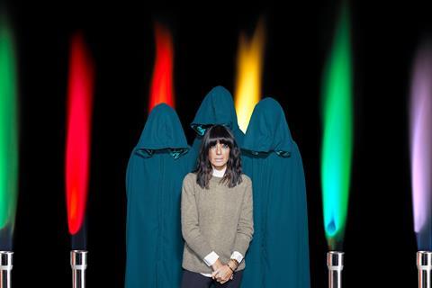 Claudia Winkleman and some cloaked hooded figures in front of some different flame colours