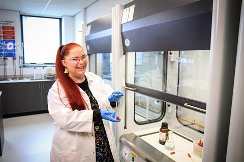 A happy teacher with a bold chemistry-theme-print dress and earrings wearing a lab coat whilst working at a fume cupboard