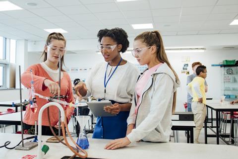 A photograph of two students and their teacher discussing a chemistry experiment, with a variety of apparatus set up in front of them