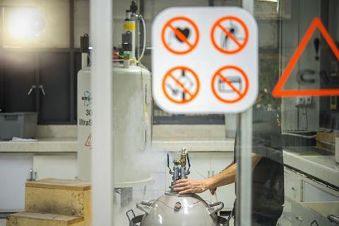 Behind a glass door with various warning signs on, a technician works with large cylinder-like lab equipment