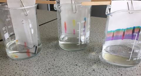 An image showing three thin layer chromatography plates; each is suspended in a measuring flask using a lolly stick and paper clip, the flasks are all on a workbench. 