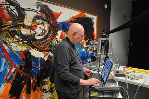 An image showing professor Jaap Boon at a computer workstation and microscope with a painting behind him