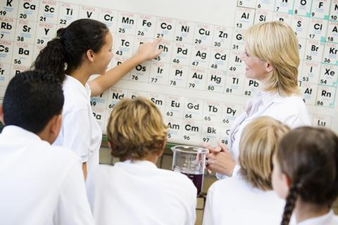 An image showing a teacher in front of a periodic table, surrounded by students, all with their backs to camera; a female student is pointing at the element cobalt.