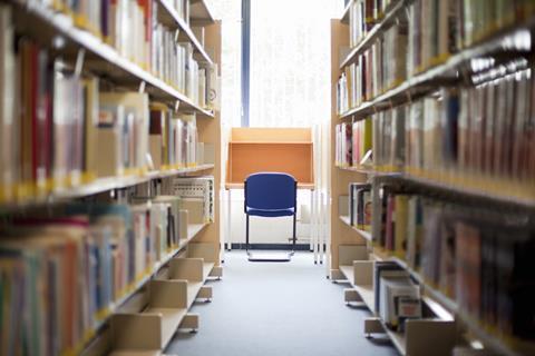 An image showing a library in a further or higher education college, two bookcases, one on either side, leading to an empty desk with a blue chair in front of a windwo