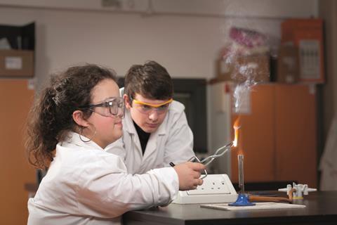 An image showing a female student burning magnesium on a Bunsen burner using tongs while a male classmate is watching the flame created. Both are wearing safety goggles