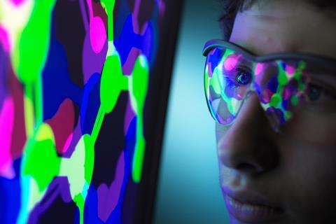 An image showing a student who is sat very close to a computer screen; a molecular structure is on the display, which reflects on his safety spectacles