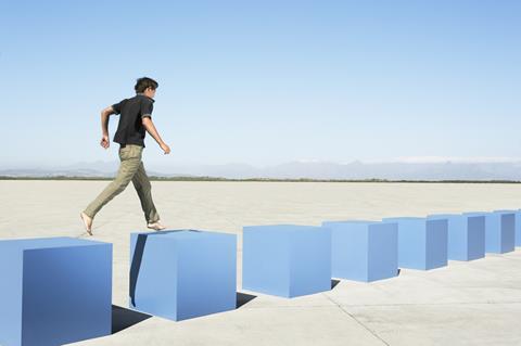 Male student barefoot walking from one blue block to another, with six more in front of him, on a concrete floor against a blue sky. Representing moving from stage to stage in an intelligent learning platform