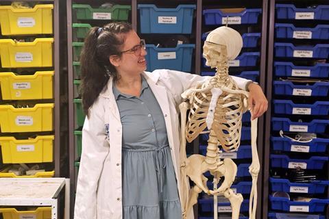 Rebecca Watkins wearing a lab coat with her arm around a teaching skeleton in a science technician's prep room