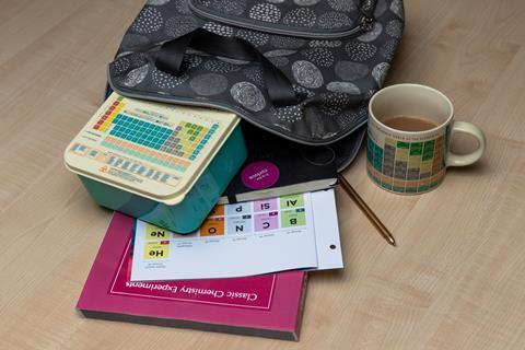 An image showing an open backpack containing a periodic table lunch box, a printed periodic table, a textbook, a notebook and a pen; a mug with the periodic table printed on it can also be seen on the table